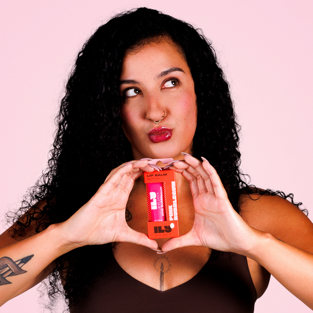 A woman with curly black hair holds a small orange product container in front of her chest, forming a heart shape with her hands. She is wearing a dark brown sports bra against a solid light pink background.